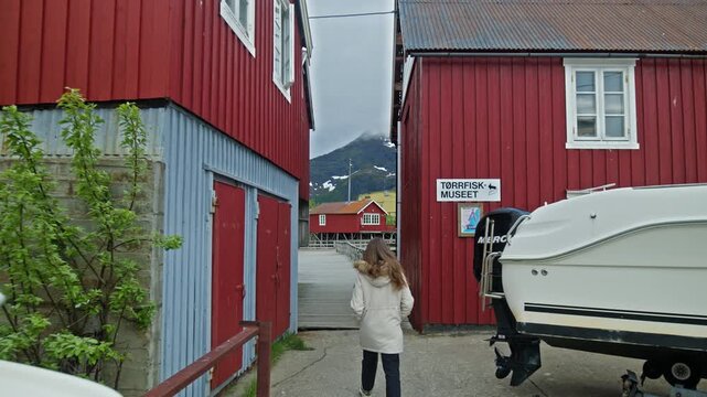 Tourist woman walkibg through a arrow passage between red wooden buildings at stockfish museum in Lofoten, Norway. Traditional fishing village architecture and cultural heritage site.