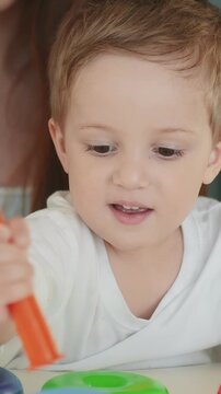 A young boy is focused on stacking colorful rings. He holds an orange stick while a woman is subtly present in the background, observing the boy's activity.