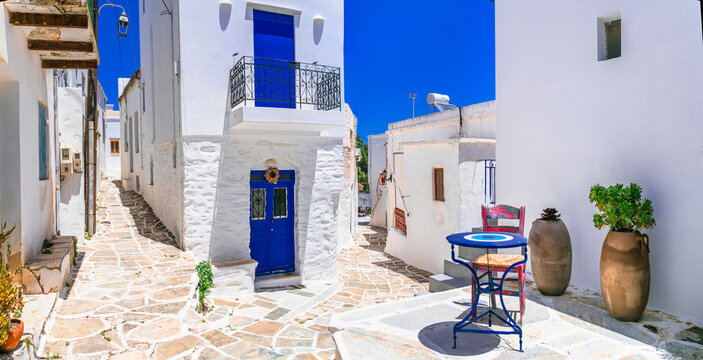 Traditional Lefkes Village Paros Greece. Empty narrow street with pink chairs with round table and ancient clay jars. Authentic Cycladic architecture white walls and blue doors Medeterranean aethetics