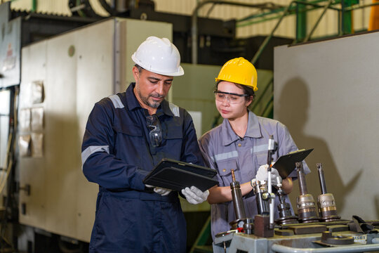 Two diverse engineers wearing hard hats looking at digital tablet discussing project plans in heavy industrial manufacturing factory plant workshop. Professional teamwork safety first concept