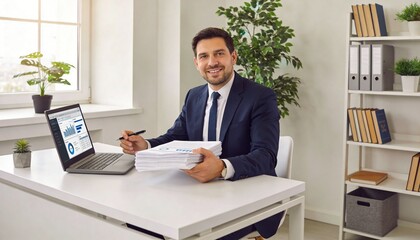 Fototapeta premium A smiling businessman sitting at a desk with a laptop and papers in a modern office