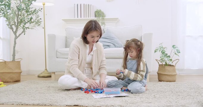 Asian single mother sitting legged, building blocks with preschool daughter, comparing colors, matching pieces with book guide, supporting child development milestone, indoor playtime livingroom.