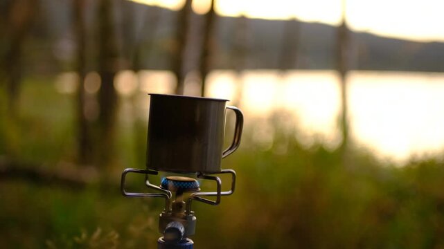 Camping mug heating on portable gas stove near lake