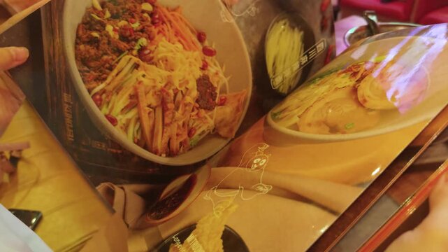 Person's hand resting on a plastic covered restaurant menu, showing various appealing illustrations of asian cuisine, vibrant noodles, and savory dishes, reflecting the process of selecting a meal