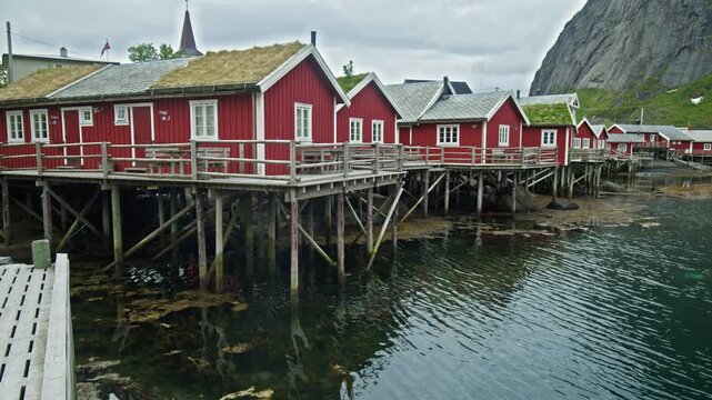 Row of red rorbuer cabins on stilts above water in Lofoten, Norway. Classic fishing village with reflections and mountain backdrop.