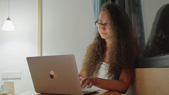 Caucasian woman typing laptop in hotel window seat, warm lamp glow, late evening work vibe, reflection in glass, focused expression, casual attire, bedside table and cozy chair visible