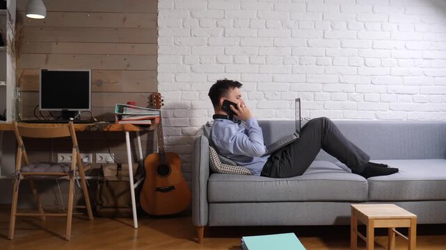Young man telecommuting from home, multitasking on his laptop while having a phone conversation, embracing a flexible work life balance from a comfortable sofa
