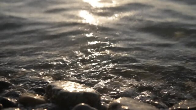 Gentle waves lap against a pebble beach with shimmering sunlight on the water.