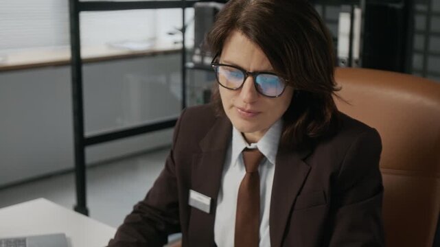 Medium close-up of focused, meticulous Caucasian female diplomat checking information in paperwork and asking questions to immigration visa applicant while working at foreign embassy
