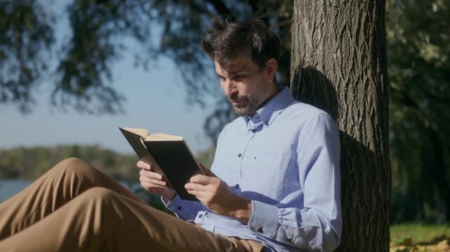 Close up of man reading book and talking while leaning on park tree