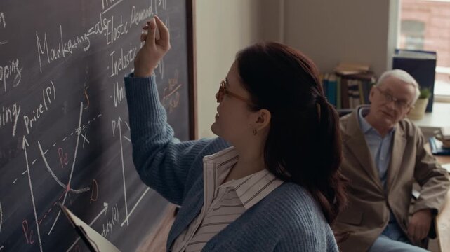 Side view shot of Caucasian female college student writing economic graph on chalkboard while senior teacher observing during lesson in modern classroom