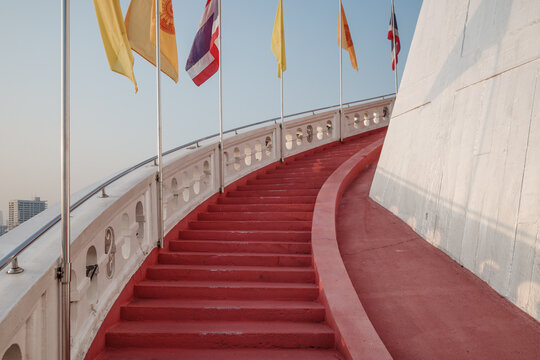 A low-angle view of a red curving staircase with white balustrades leading up the Golden Mount at Wat Saket in Thailand, decorated with waving Thai and Buddhist flags against a clear sky.