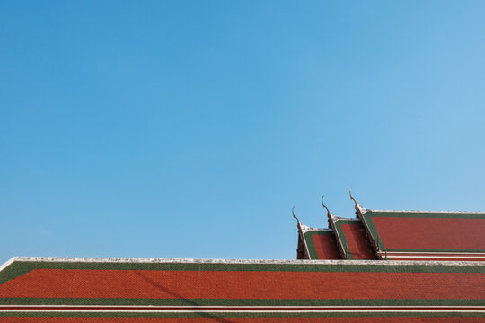 Traditional Thai temple roof with red and green tiles and golden chofah ornaments against a cloudless blue sky