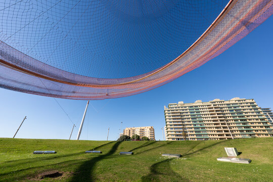 Modern apartment buildings with large net canopy above green grass park under clear blue sky.
