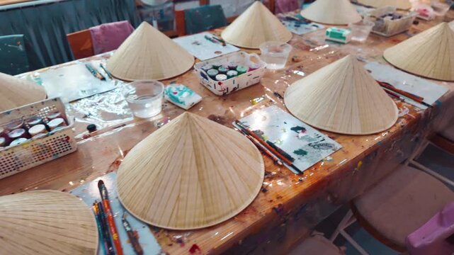 Non la hat painting workshop with brushes palettes and colorful paints on wooden table, Traditional Vietnam craft activity painting conical hats in creative art studio setting 4k