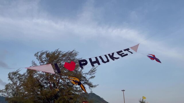 Colorful &ldquo;I love Phuket&rdquo; banner waving in the wind during lively Karon kite festival with kites flying in the sky
