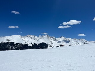 Fototapeta premium Wide view of a snow-covered mountain range and ski slope under a deep blue sky with scattered white clouds on a clear winter day.