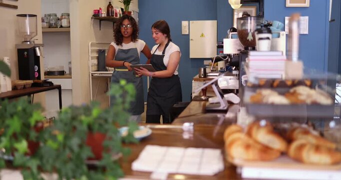 Two diverse female employees working at a local coffee shop, using a digital tablet to manage orders and inventory, showcasing teamwork and modern small business operations