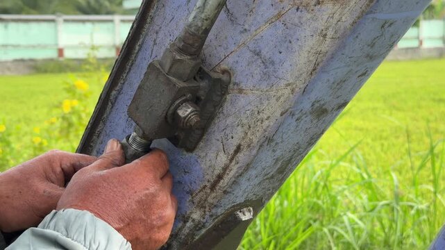 A worker tightens a bolted metal connection on an excavator mechanism using a wrench while performing service work beside a green agricultural field outdoors. Engineering work.