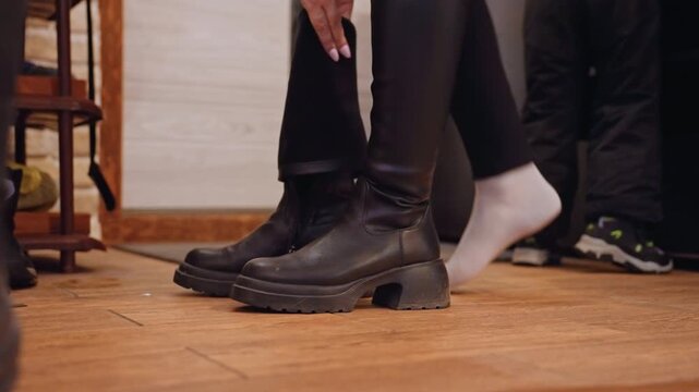 Mom helping son put on boots in hallway with wooden floor, black leather boots mother kneeling to zip and adjust boot while child stands in sock, warm domestic atmosphere, tender caregiving