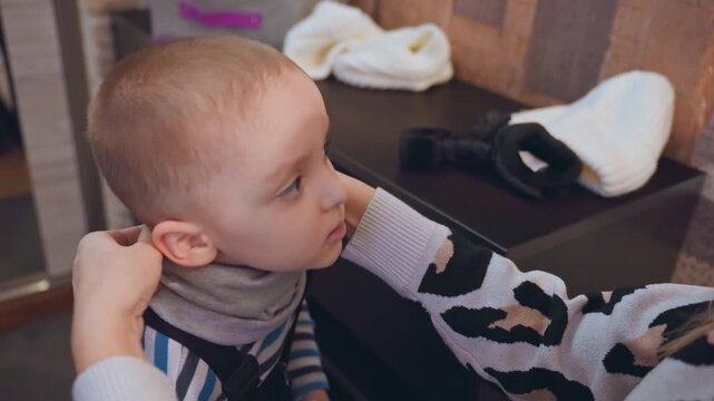 White mom puts hat on son, adjusts scarf and playfully covers eyes with sock young boy winces, frowns then calms as mom removes covering, gentle hallway morning routine with overalls, knit beanie