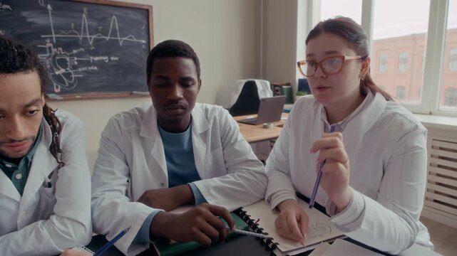Tracking tilt down closeup shot of diverse medical students in lab coats discussing female reproductive system anatomical model with uterus and ovaries at desk in classroom