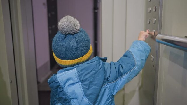 Back view toddler reaching elevator rail, white son in blue coat and knit beanie with pompom, small hand gripping metal handrail while looking into lit hallway, mom nearby, cozy winter outing vibe