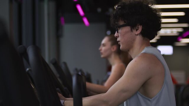Side view of young man and woman doing intensive cardio, using elliptical trainers in modern gym