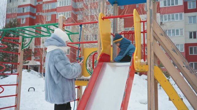 White mom helps son on slide bundled winter clothing urban apartment backdrop colorful plastic slide caring encouragement as child sits at top gentle supervision and warm interaction