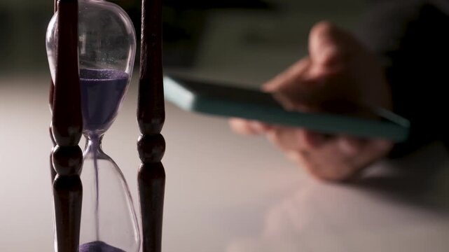 Person checking smartphone next to hourglass on desk, concept of deadline and ti