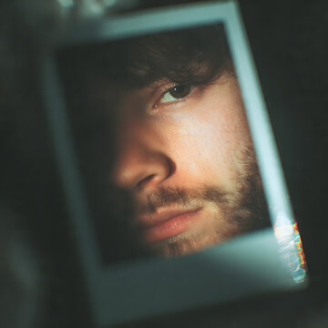 A close-up photograph of a man's face, with a Polaroid effect, focusing on his intense, expressive eyes and slight beard.