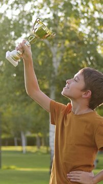 Happy child holding trophy and celebrating victory in park at sunset. Childhood dreams, aspiration and future success. Joyful boy imagines becoming winner. Inspiration, motivation and dream. Vertical.