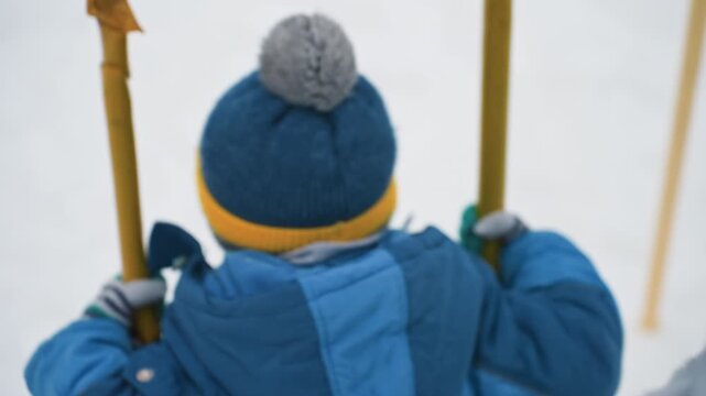 Son back on winter swing closeup, bundled child in blue coat and pompom hat, rope and grip details, offscreen mother hand gently steadying, quiet reflective mood, fabric texture and soft breath