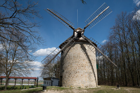 Historic stone windmill with wooden blades and a shingle roof standing on a hill under a bright blue sky with white clouds, Holic, Slovakia