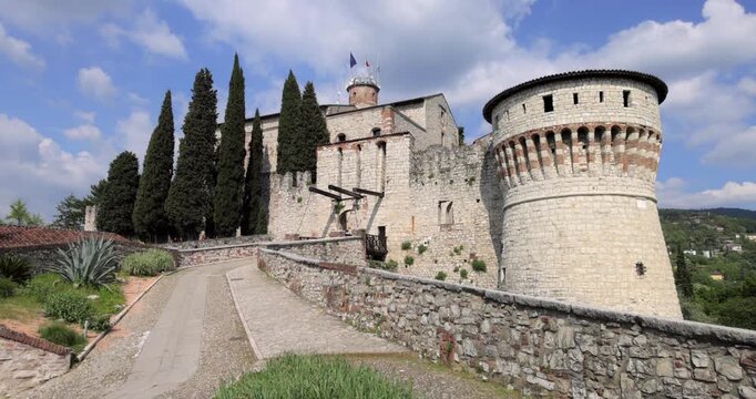 Castle of Brescia fortress tower with fluttering flags in Brescia Italy, wide static view shows sunlit stone ramparts and cypress trees as Italian tricolor and European Union banners wave