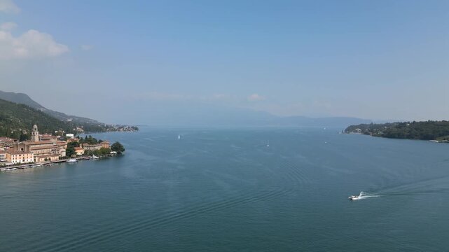 Drone curved approach over Lake Garda to Salo old town, drone glides along the shore highlighting Santa Maria Annunziata cathedral, marina and rippling water with sailboats on a bright day