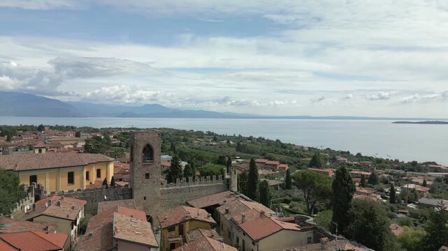 Drone yaw orbit above castle of Moniga del Garda in Italy, slow panoramic sweep reveals town rooftops cypress trees and Lake Garda with distant mountains