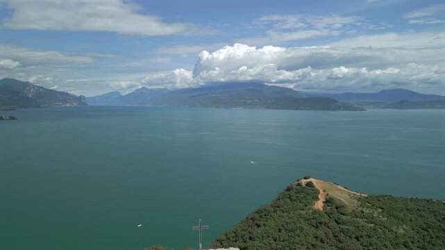 High drone pullback over Rocca di Manerba del Garda ruins, revealing forested promontory and Lake Garda panorama in bright sunny summer daylight