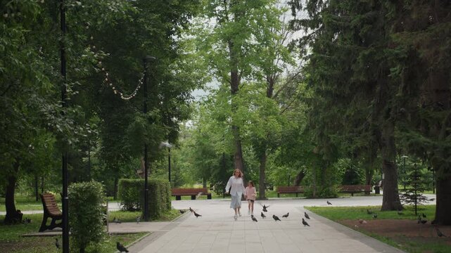 Mom and daughter running through park, mother encouraging child along treelined path, pigeons flushing into sky, motion blur and summer breeze, active playful bond, candid outdoor exercise, smiling