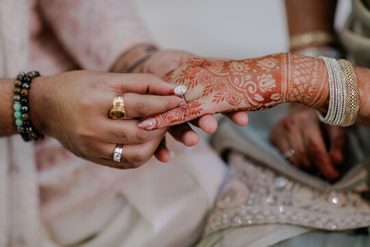 Close-up shot of a groom gently placing a beautiful engagement ring onto the bride's hand adorned with intricate henna designs, symbolizing commitment
