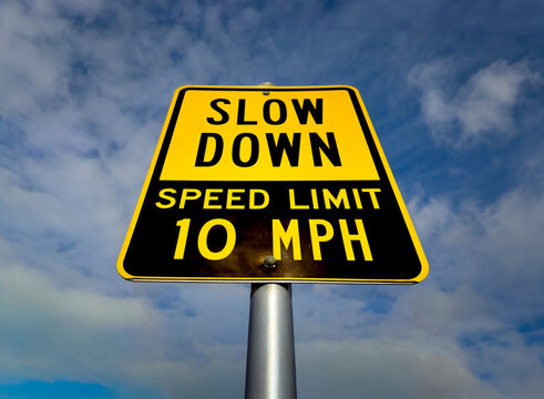 A yellow sign in a parking lot stating Slow Down Speed Limit 10 MPH against a blue sky