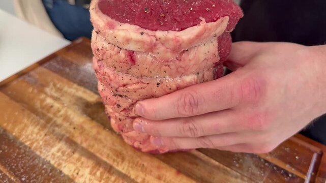 Closeup of a man&rsquo;s hands turning a raw topside joint of beef on a wooden board in a kitchen, while black pepper seasoning is shaken all over the uncooked meat.