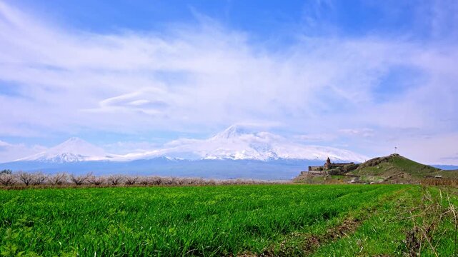 Vast green fields stretch toward snow-capped Ararat mountain under blue skies. Khor Virap Monastery