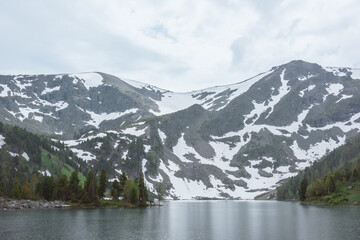 Obraz premium Mountain lake in rainy weather. Alpine lake between green hills with conifer forest with view to high rocky snowy mountain wall under gloomy cloudy sky. Circles of heavy rain on gray water surface.