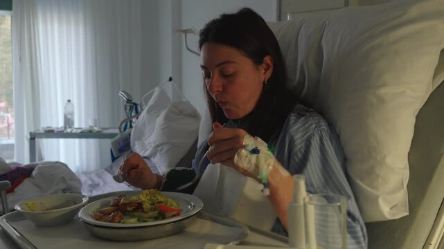 Female patient in hospital bed eating carefully with intravenous catheter in hand, lifting food to mouth, expressing weakness, recovery effort and daily routine during treatment