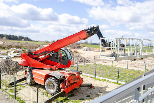 Red telescopic handler lifting platform on construction site with green grass and scattered building materials under a blue sky with clouds in the background