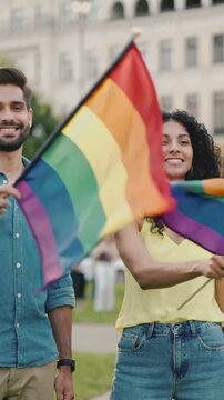 Vertical video: Flag shifting couple in yellow top and denim waving rainbow flags for pride at park