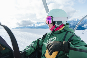 Happy female snowboarder resting on a ski lift in high mountains during winter season. Young woman in helmet and mirrored goggles enjoying snowboarding, freedom and active lifestyle. Bright sunny day  © Annatamila
