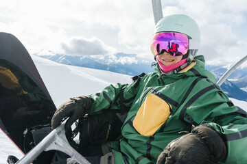 Happy female snowboarder resting on a ski lift in high mountains during winter season. Young woman in helmet and mirrored goggles enjoying snowboarding, freedom and active lifestyle. Bright sunny day  © Annatamila