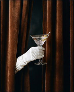 A hand wearing a vintage white glove holding a martini glass with olives, emerging from brown velvet curtains